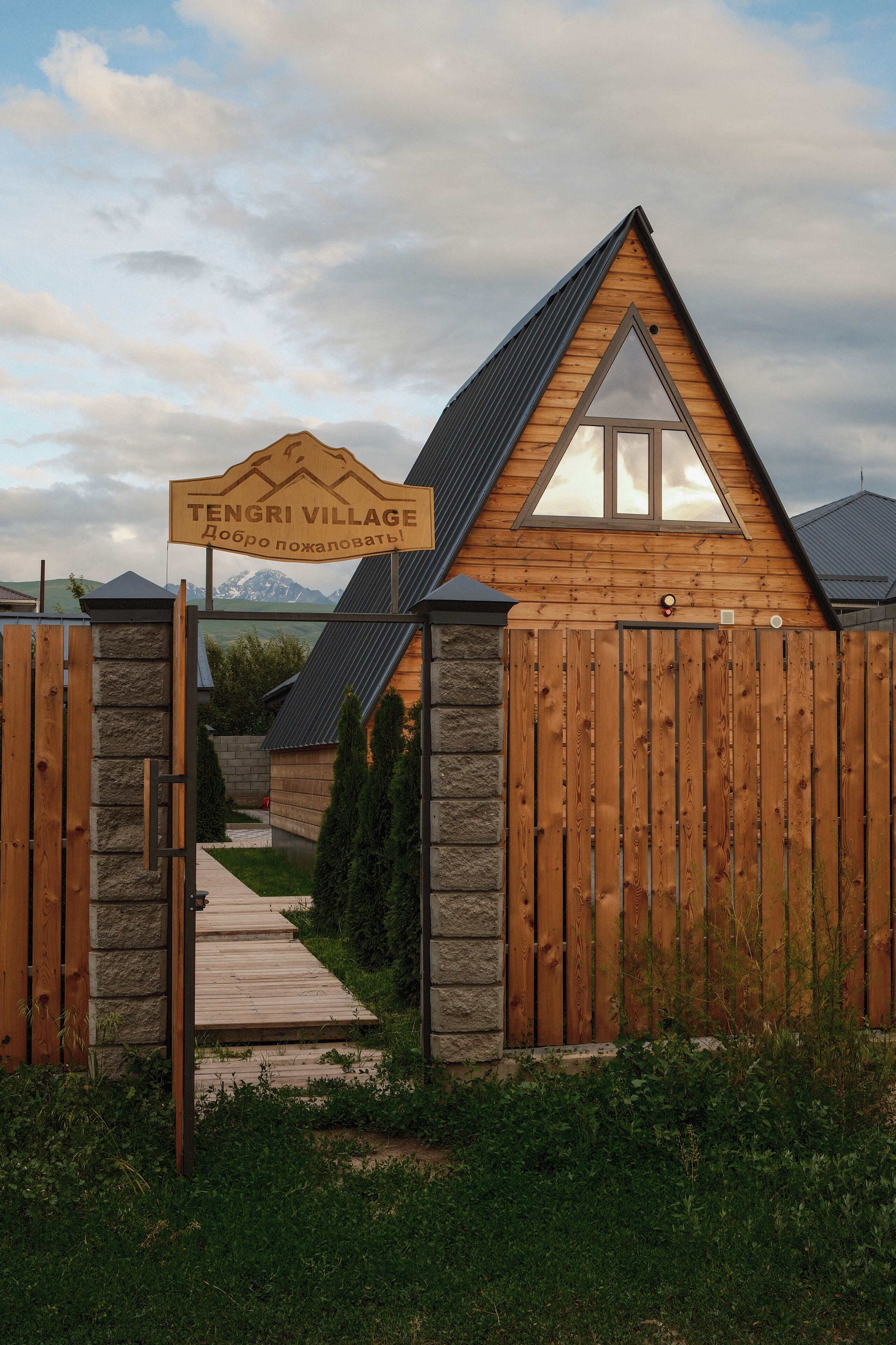A wooden cabin with a stone wall entrance, a wooden gate, and a sign reading 'Tengri Village.'