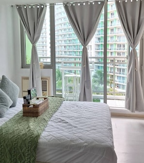 A bedroom with a bed, a window with curtains, a view of a high-rise building, and a small table with a basket.