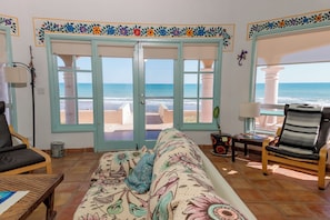 Ein Zimmer mit einer Couch, einem Sessel, einer Lampe und Blick auf das Meer durch eine Tür.