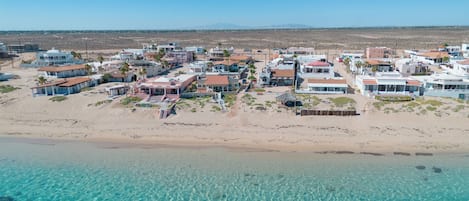 An aerial view of a coastal resort with beachfront villas, clear turquoise waters, and a desert landscape in the background.