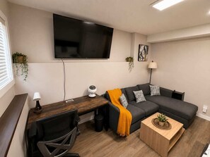 A modern living room with a flat-screen TV, a sofa, a coffee table, a desk with a lamp, and a chair.