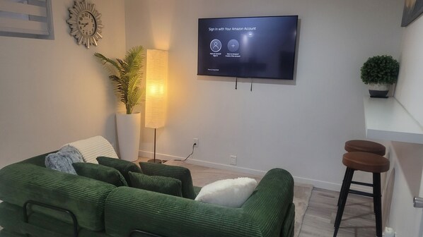 Ein Wohnzimmer mit einem Flachbildfernseher, der einen Anmeldebildschirm anzeigt, einer großen Topfpflanze, einer Stehlampe, einem grünen Sofa und einem kleinen Hocker.