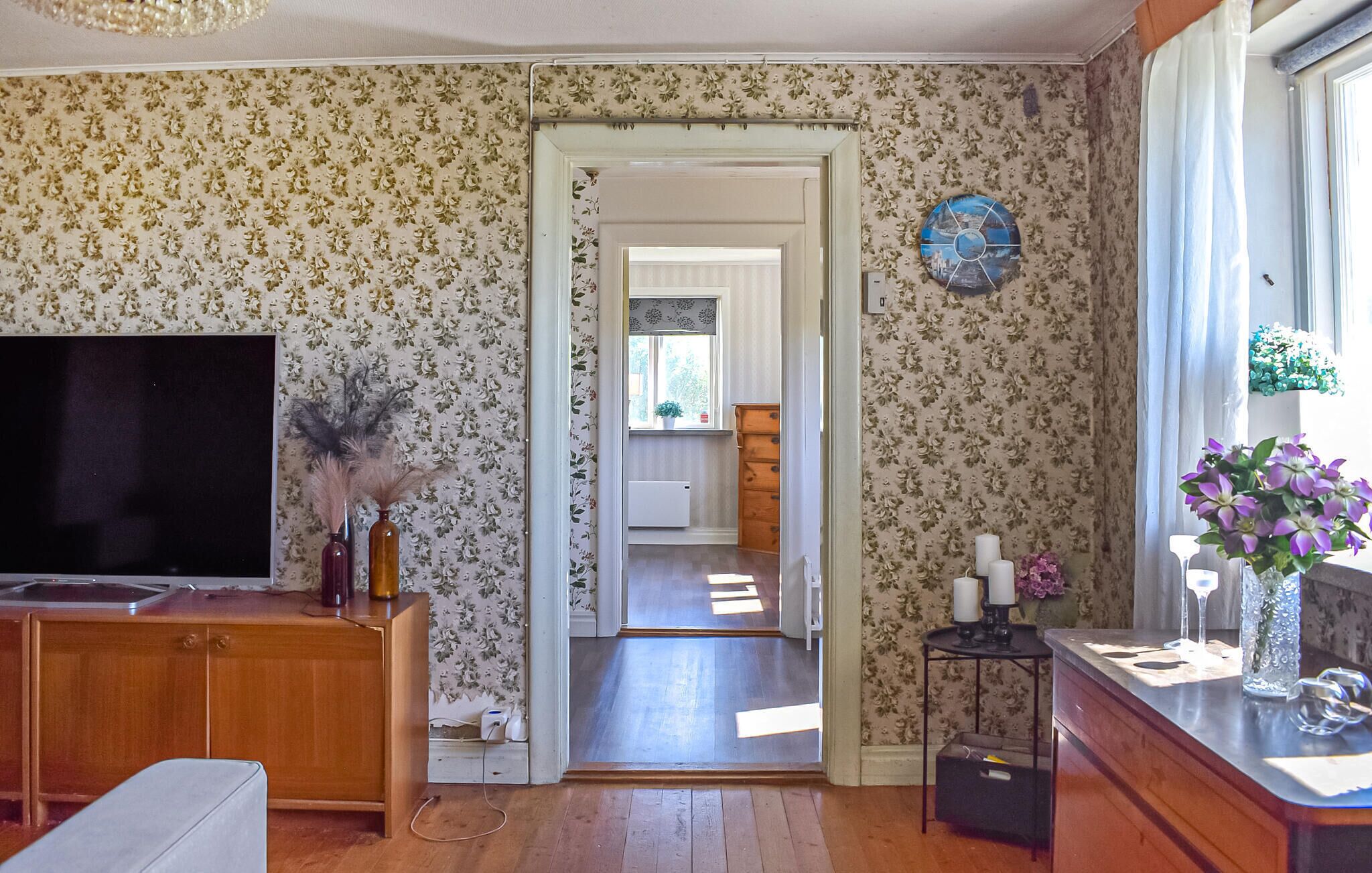A room with floral wallpaper, a television, a wooden cabinet, a small table with candles, and a window with a view.