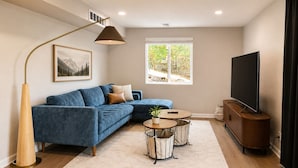 A living room with a blue sofa, a coffee table, a lamp, a TV, and a window with a view of greenery. - 1,100 sf Spacious Cozy 2BR Near Denver DT, Red Rocks, and Rockies! (Arvada)