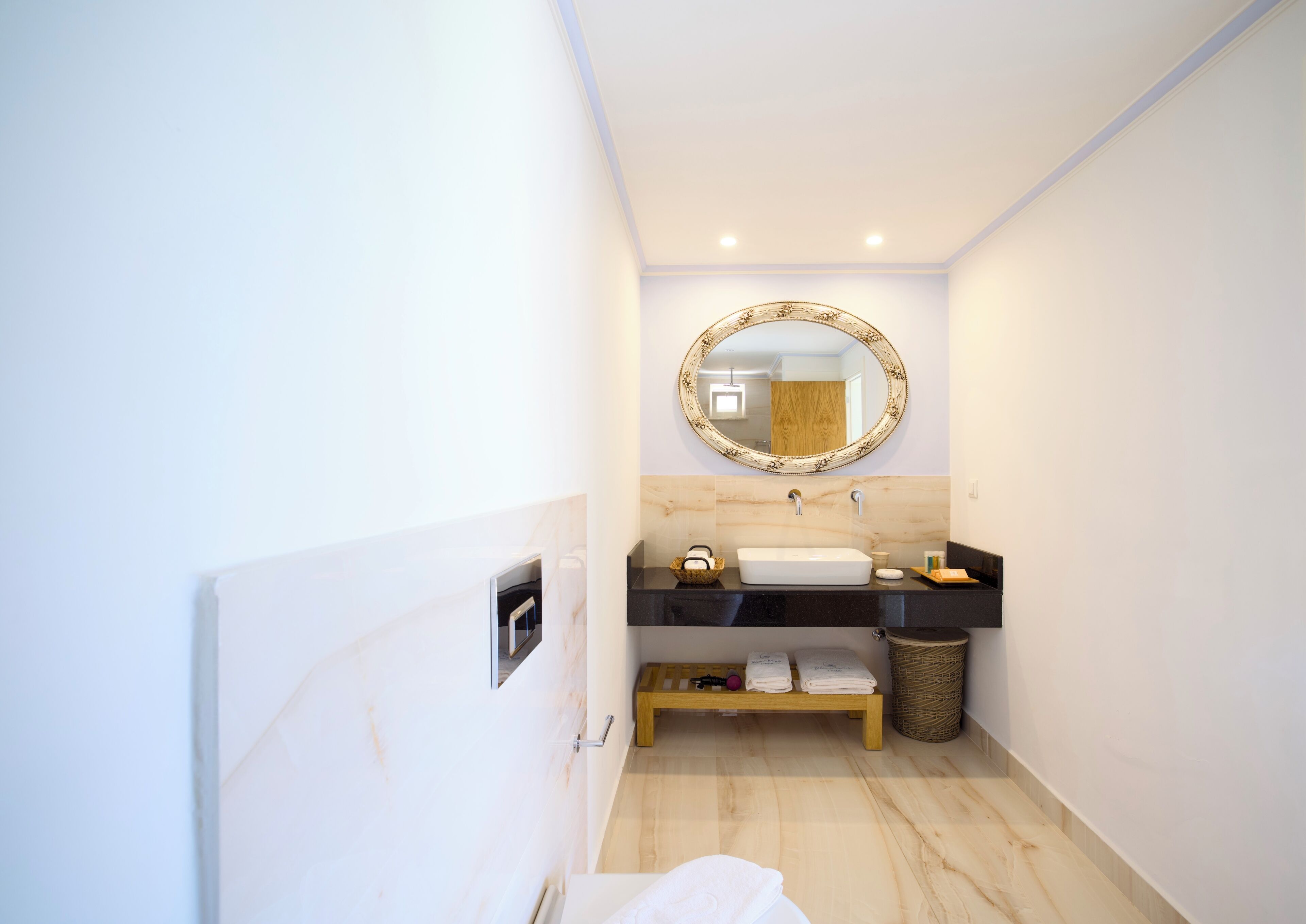 A modern bathroom with a marble sink, a round mirror, and a wooden bench.