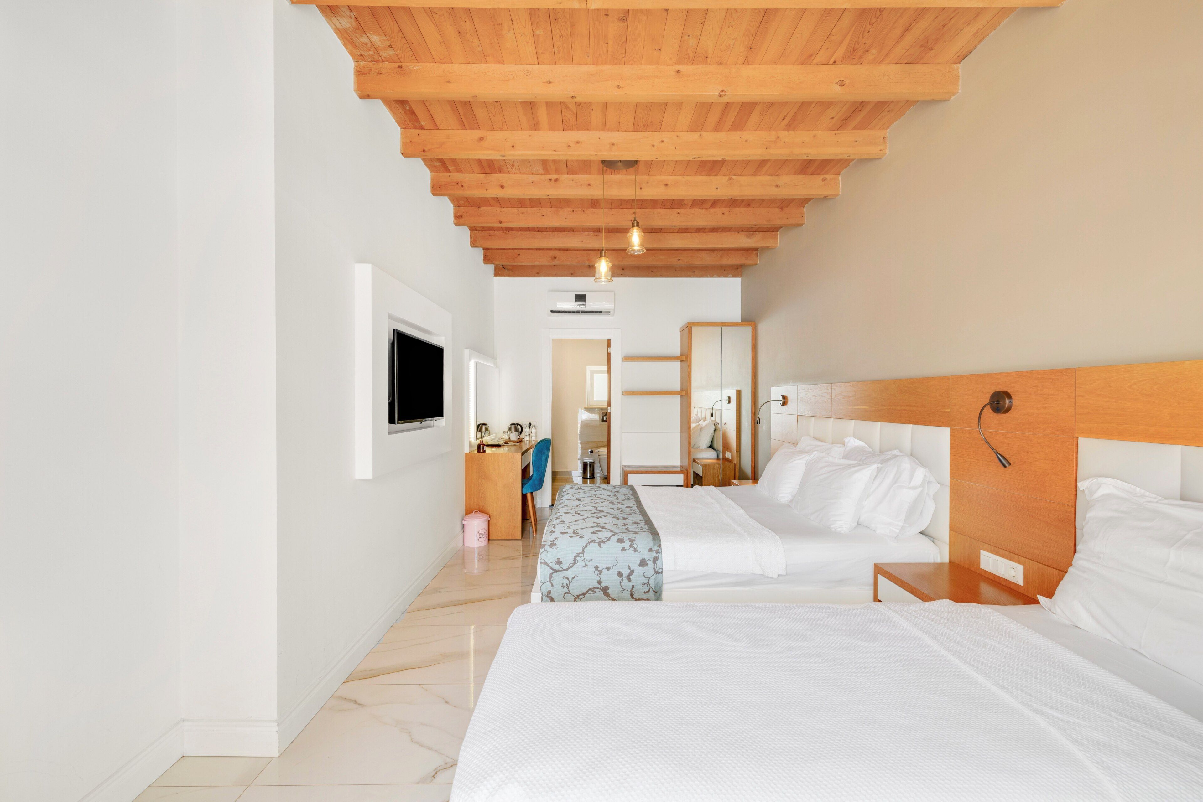 A hotel room with two beds, a wooden ceiling, a TV, a desk, and a mirror.