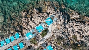 An aerial view of a coastal hotel with multiple blue-roofed structures, a wooden deck, and clear turquoise water.