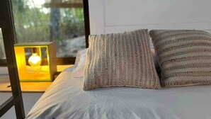 Ein Bett mit zwei strukturierten Kissen, ein Nachttisch mit Lampe und Blick auf Bäume durch ein Fenster.