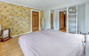 Ein Schlafzimmer mit einem Bett, Holzboden, blumigem Wandpapier und einer Tür zu einem anderen Raum.