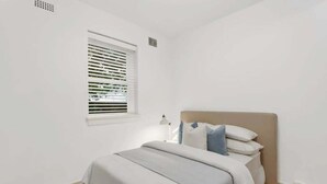 Un dormitorio con cama, ventana con persianas, lámpara de noche y una pequeña alfombra.