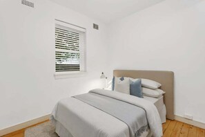 Ein Schlafzimmer mit Bett, einem Fenster mit Jalousien, einer Nachttischlampe und einem kleinen Teppich.