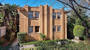 Un edificio de dos pisos con fachada simétrica, numerosas ventanas y un jardín cuidado con diversas plantas y arbustos podados.