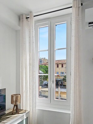 Une pièce avec une grande fenêtre, des rideaux blancs et une petite table avec une lampe.