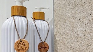 Two white bottles with wooden caps and labels hanging from a white shelf against a textured wall.