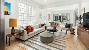 A spacious living room with a sofa, ottoman, and coffee table, large windows, and a television. - Ultralux 5BDR Beachfront Home in Elwood (Elwood)