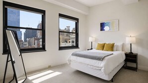 A bedroom with a bed, two bedside lamps, a painting, and a city view through large windows. - 2 BR Iconic Detroit High-Rise Loft (Detroit)