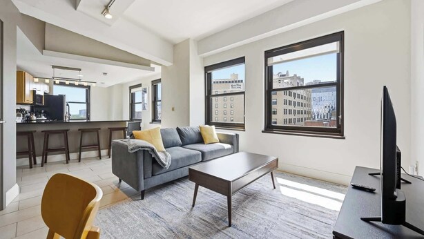 A modern living room with a sofa, coffee table, and TV, large windows with city views, and a kitchen area. - 2 BR Iconic Detroit High-Rise Loft (Detroit)