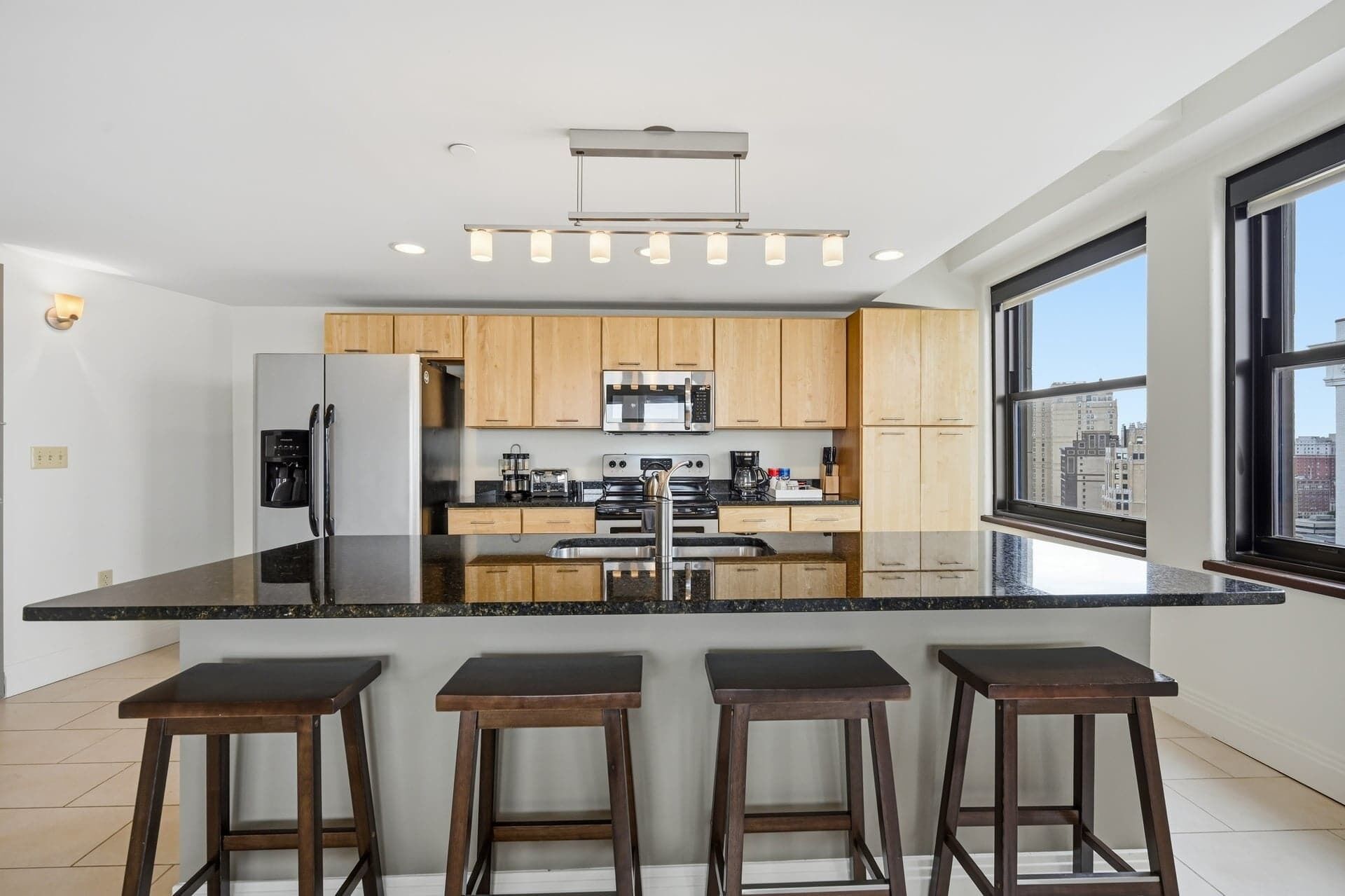 A modern kitchen with a central island, wooden cabinets, stainless steel appliances, and a large window offering a city view.