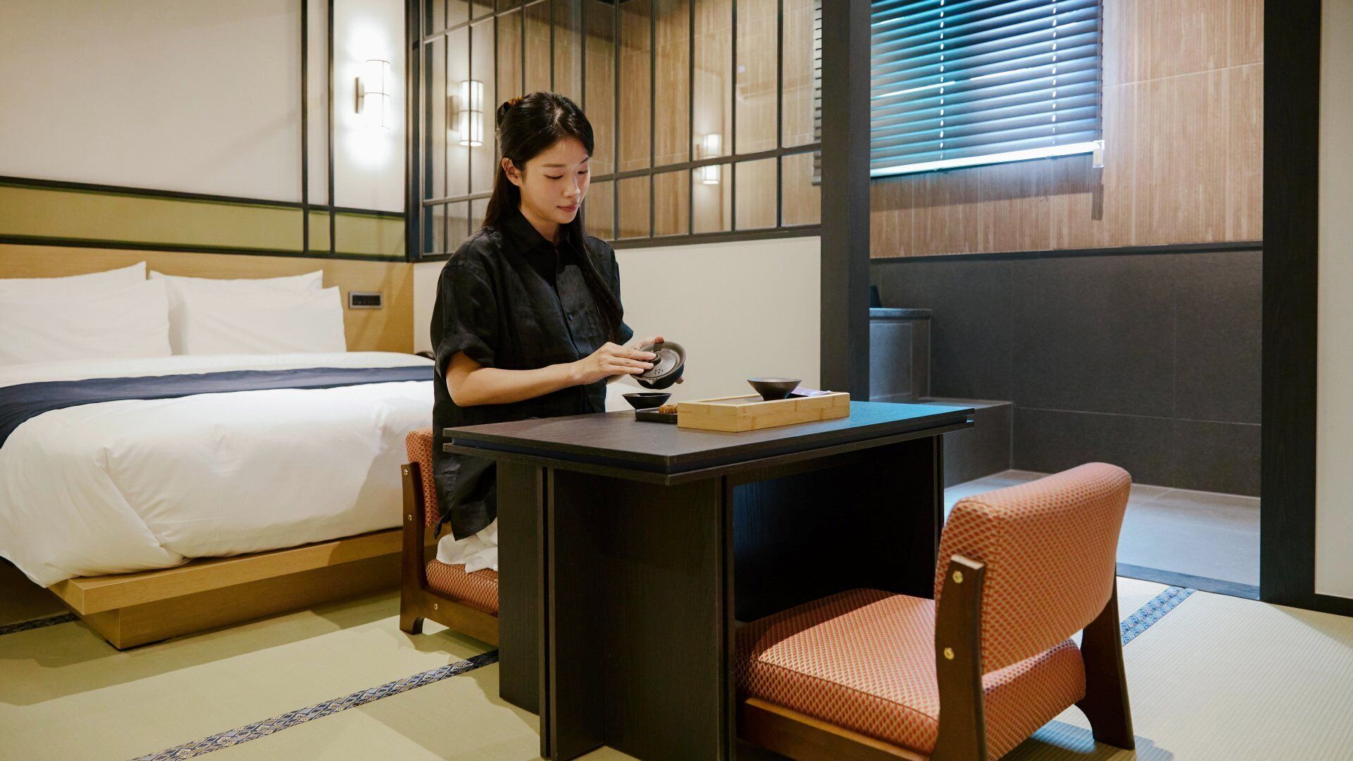 A traditional Japanese room with tatami flooring, a low table, a chair, a bed with white bedding, and sliding doors.