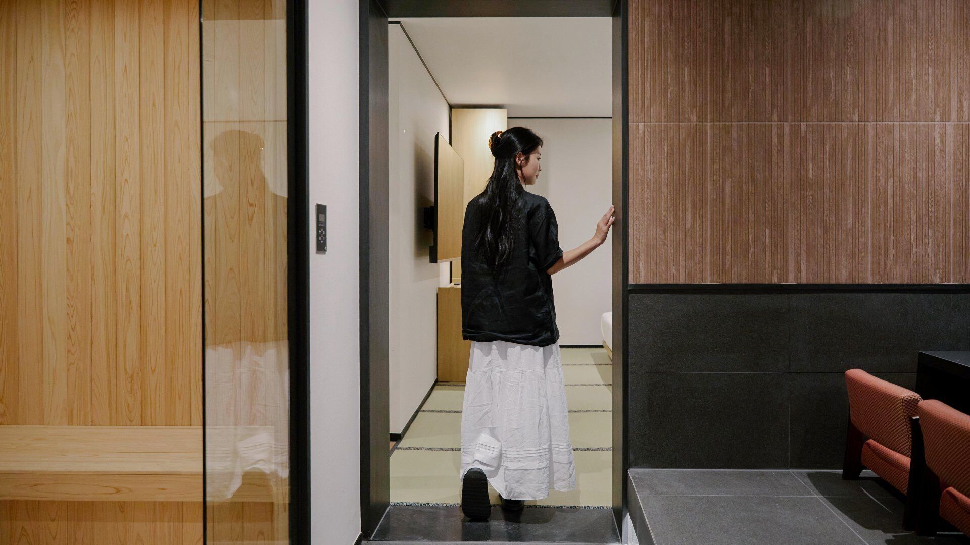 A person entering a room with a wooden door, a black wall, and a red bench.