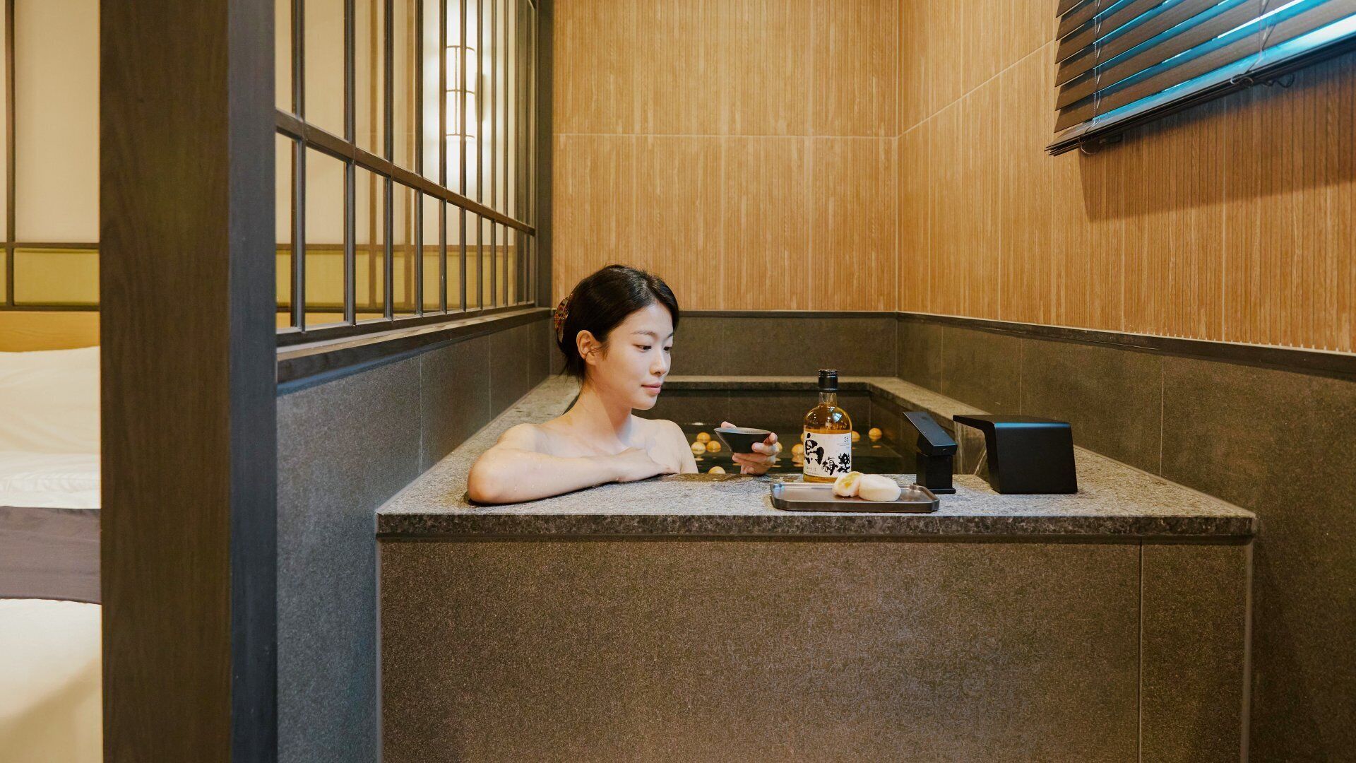 Una persona relajándose en un entorno tipo spa, con una bañera, una botella de loción y un cuenco.
