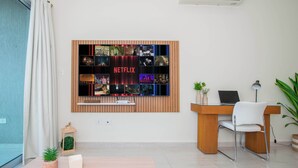 Een moderne woonkamer met een aan de muur bevestigde televisie waarop een Netflix-interface zichtbaar is, een houten bureau, een witte stoel, een kleine plant en een lamp.