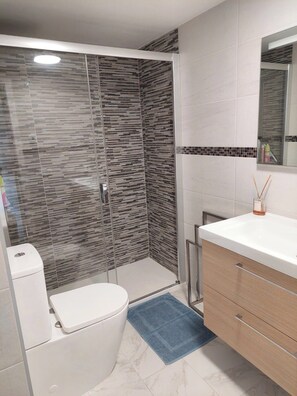 Une salle de bain moderne avec une douche à parois de verre, des toilettes blanches, un meuble-lavabo en bois avec un miroir et un tapis bleu.