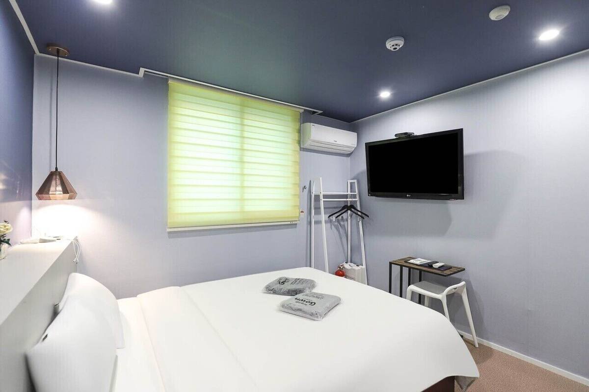 A bedroom with a bed, a TV mounted on the wall, a window with blinds, and a bedside table.