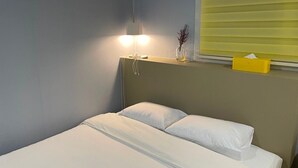 Una cama individual con ropa de cama blanca, una lámpara de noche y una ventana con persianas.