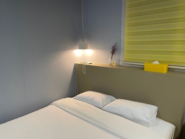 Una cama individual con ropa de cama blanca, una lámpara de noche y una ventana con persianas.