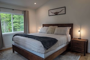 Ein Schlafzimmer mit einem großen Bett, einem Nachttisch mit Lampe, einem gerahmten Bild an der Wand und einem Fenster mit Vorhängen.