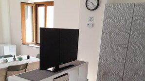 A room with a television on a white cabinet, a clock on the wall, and a window with a view.