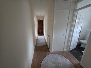Een smalle gang met een houten vloer, een witte deur, en een zichtbare badkamer met toilet en wastafel.