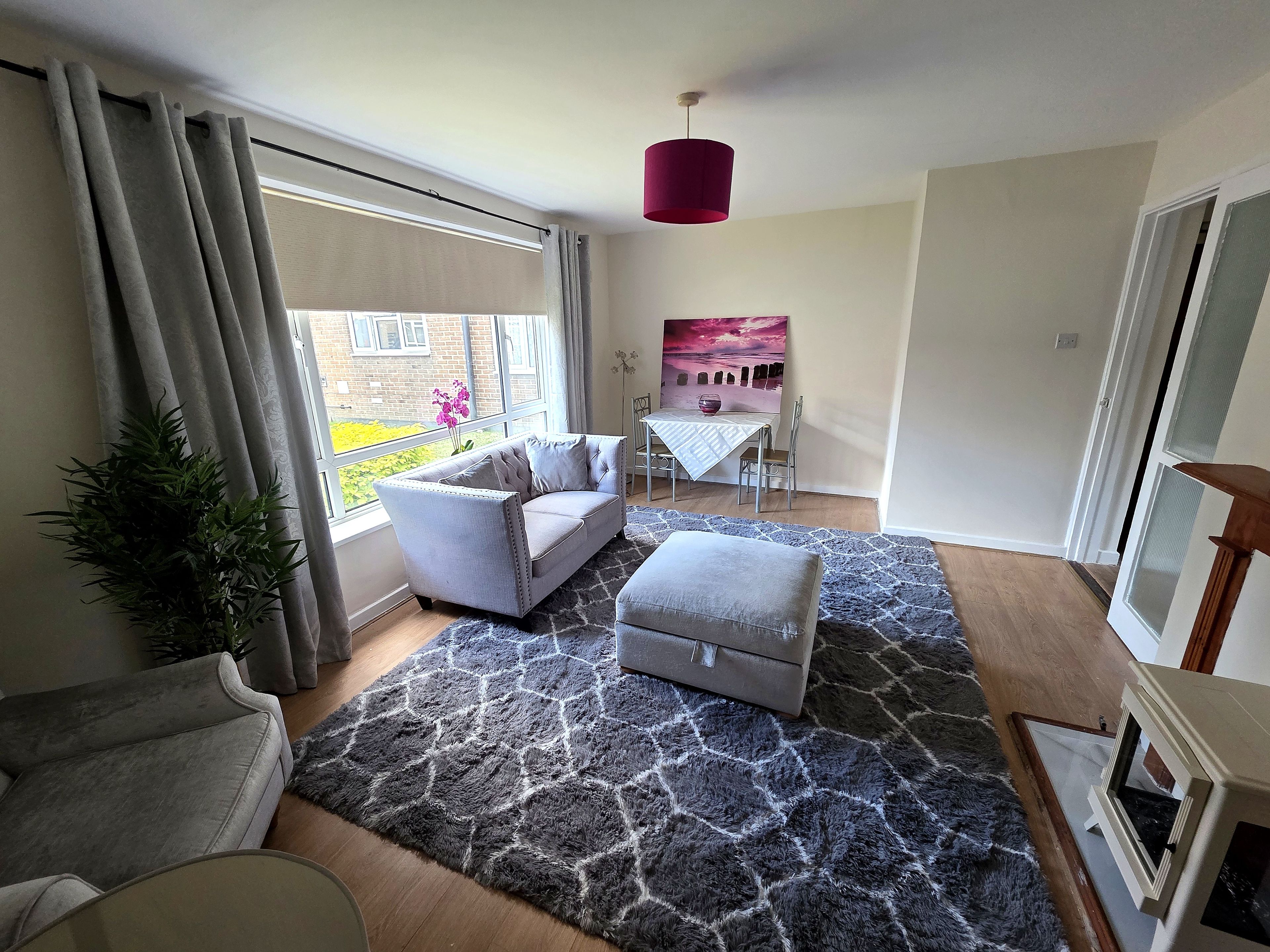 A modern living room with a grey sofa, a matching ottoman, a patterned rug, and a dining area with a table and chairs.
