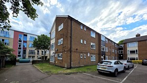 A multi-story residential building with a brick facade, multiple windows, and a parking area with cars. - Modern 2BDR Flat Near Stratford and Central London (Greater London)