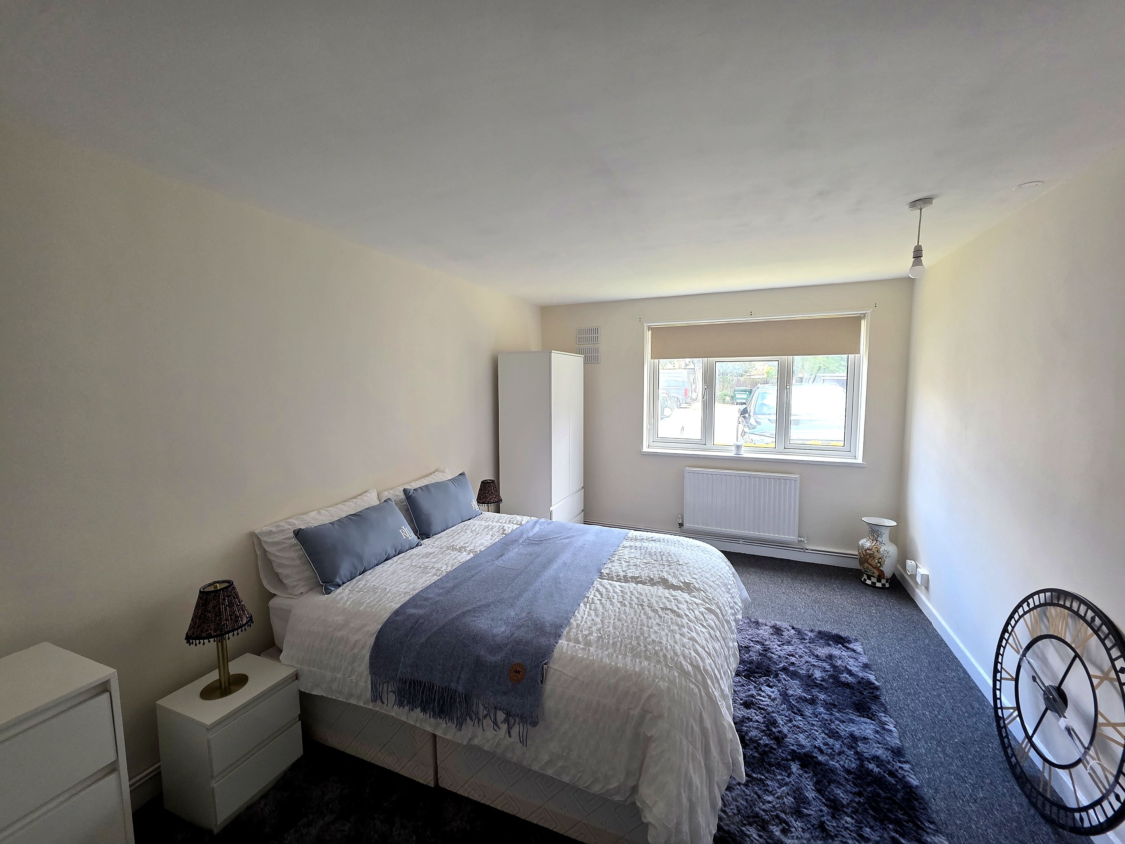 A bedroom with a bed, bedside table, lamp, radiator, window, and a wall clock.