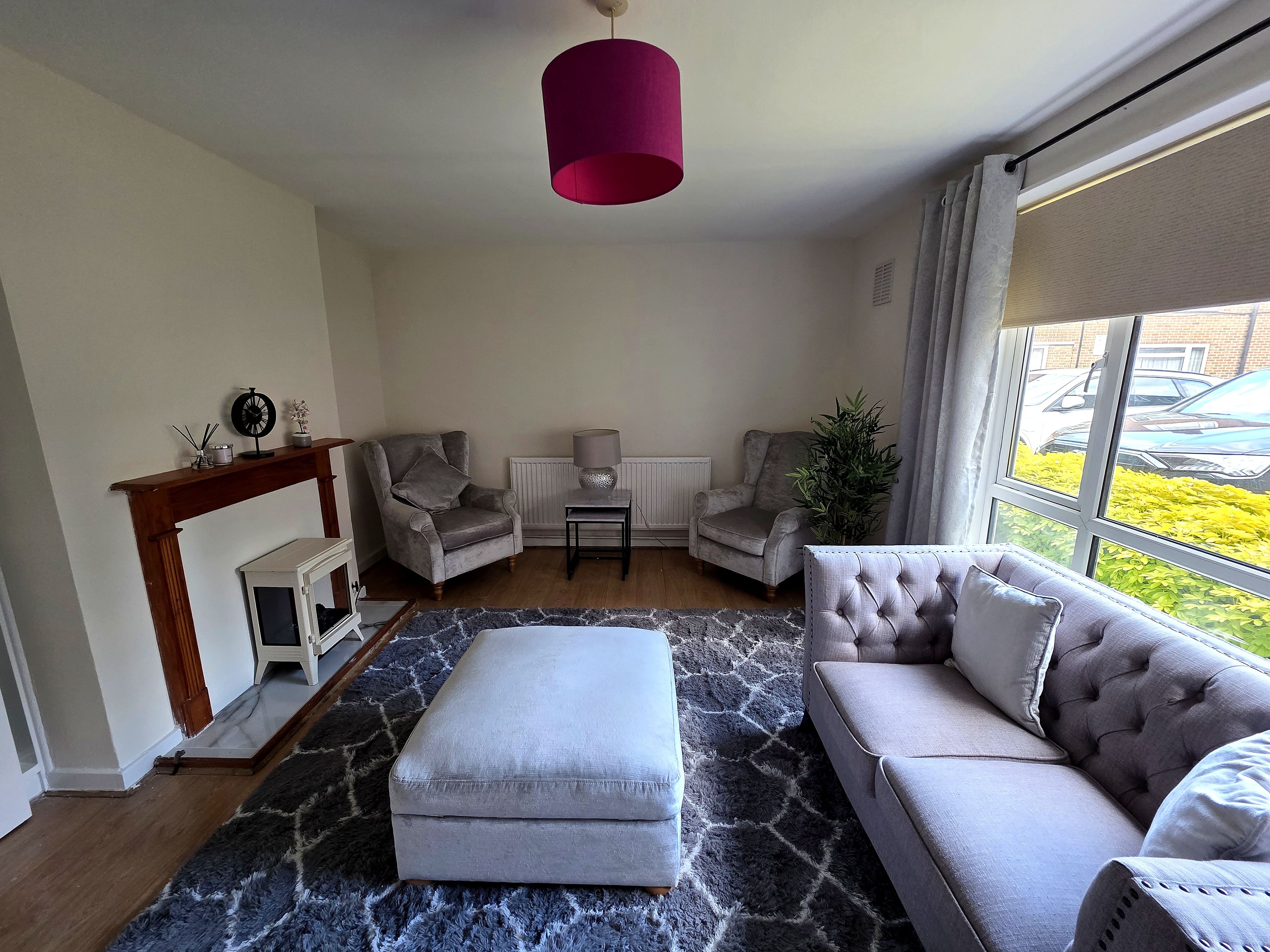 Ein Wohnzimmer mit grauem Sofa, passendem Hocker, zwei Sesseln, einem kleinen Tisch, einer Stehlampe und einem großen Fenster mit Blick auf Autos und Grünflächen.