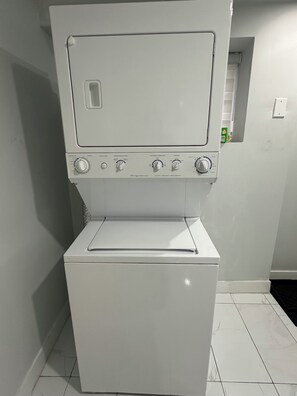 A white washing machine with control knobs and a door, placed in a room with white tiled flooring and a small window. - Newly renovated private basement Suite - central hub (Vancouver)