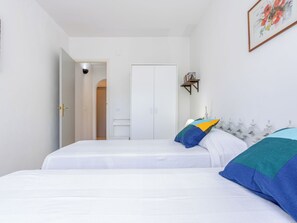 A bedroom with two beds, a white wardrobe, a door leading to another room, and a small shelf with books. - Mestral by Interhome (Palau Savardera)