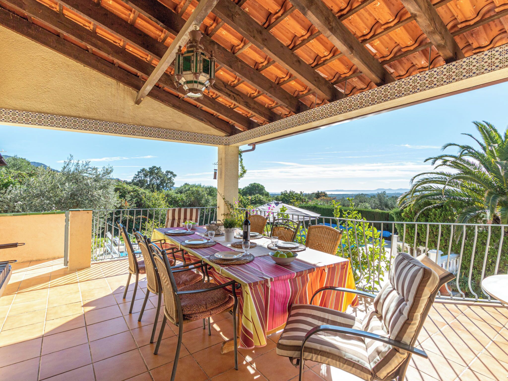 An outdoor patio with a table set for dining, surrounded by chairs, under a wooden ceiling with a patterned border.