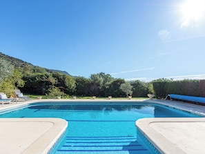 A swimming pool with clear blue water, surrounded by lounge chairs and potted plants, with a mountain in the background. - Mestral by Interhome (Palau Savardera)