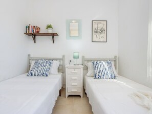 A twin room with two beds, a bedside table with a lamp, a small shelf with books and a plant, a framed picture on the wall, and a mirror above the bed. - Mestral by Interhome (Palau Savardera)