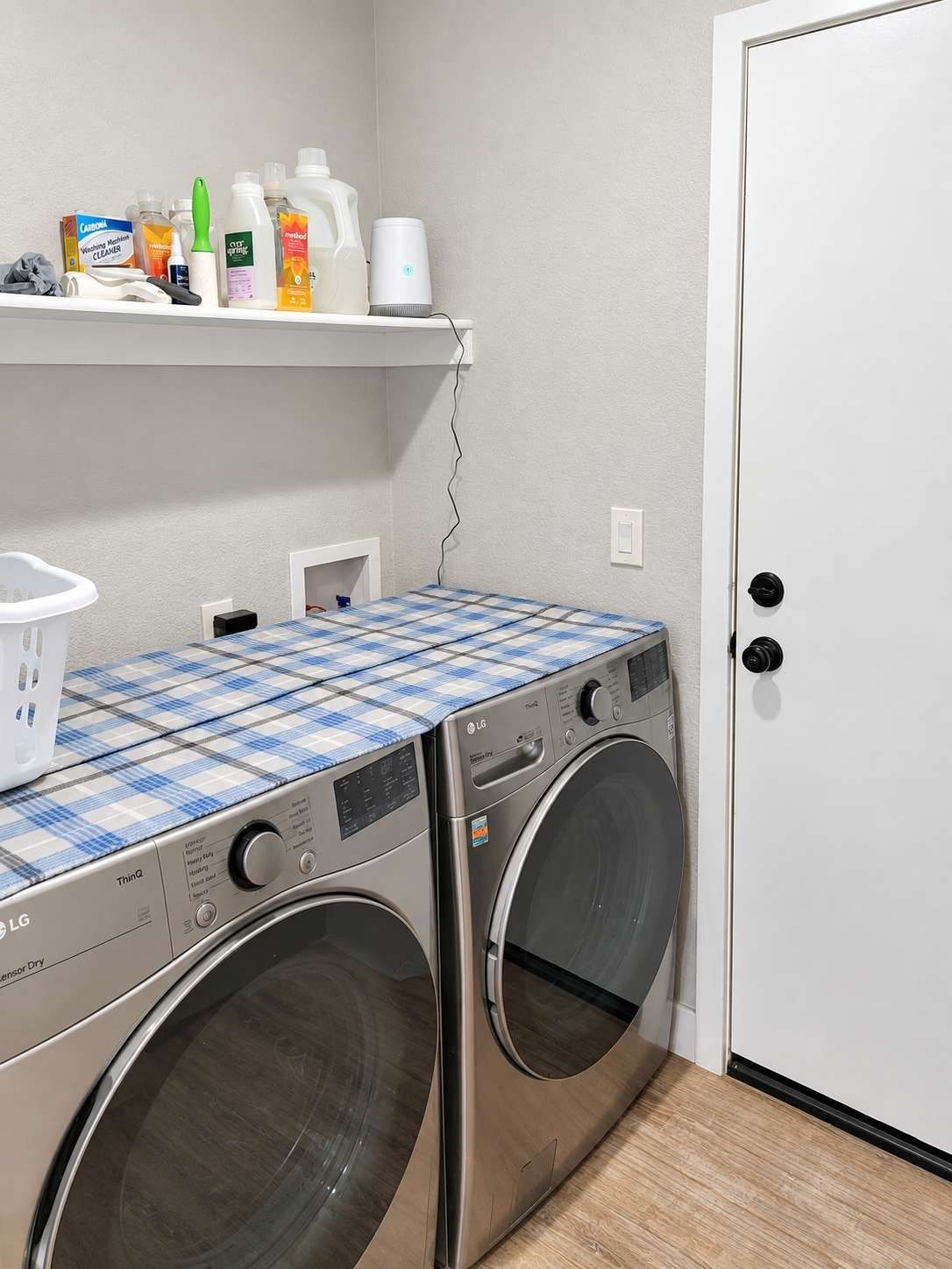 A laundry area with two front-loading LG washers, a shelf with cleaning supplies, and a white door.