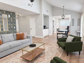 A spacious living area with a sofa, a coffee table, and a green armchair. There is a kitchen in the background with a dining table and chairs. - Chandler 4BR | Private Pool | Pet Friendly (Chandler)