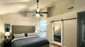 Une chambre à coucher avec une tête de lit capitonnée, un lit avec une literie aux couleurs sombres, un ventilateur de plafond, un dressing et un tapis à motifs.
