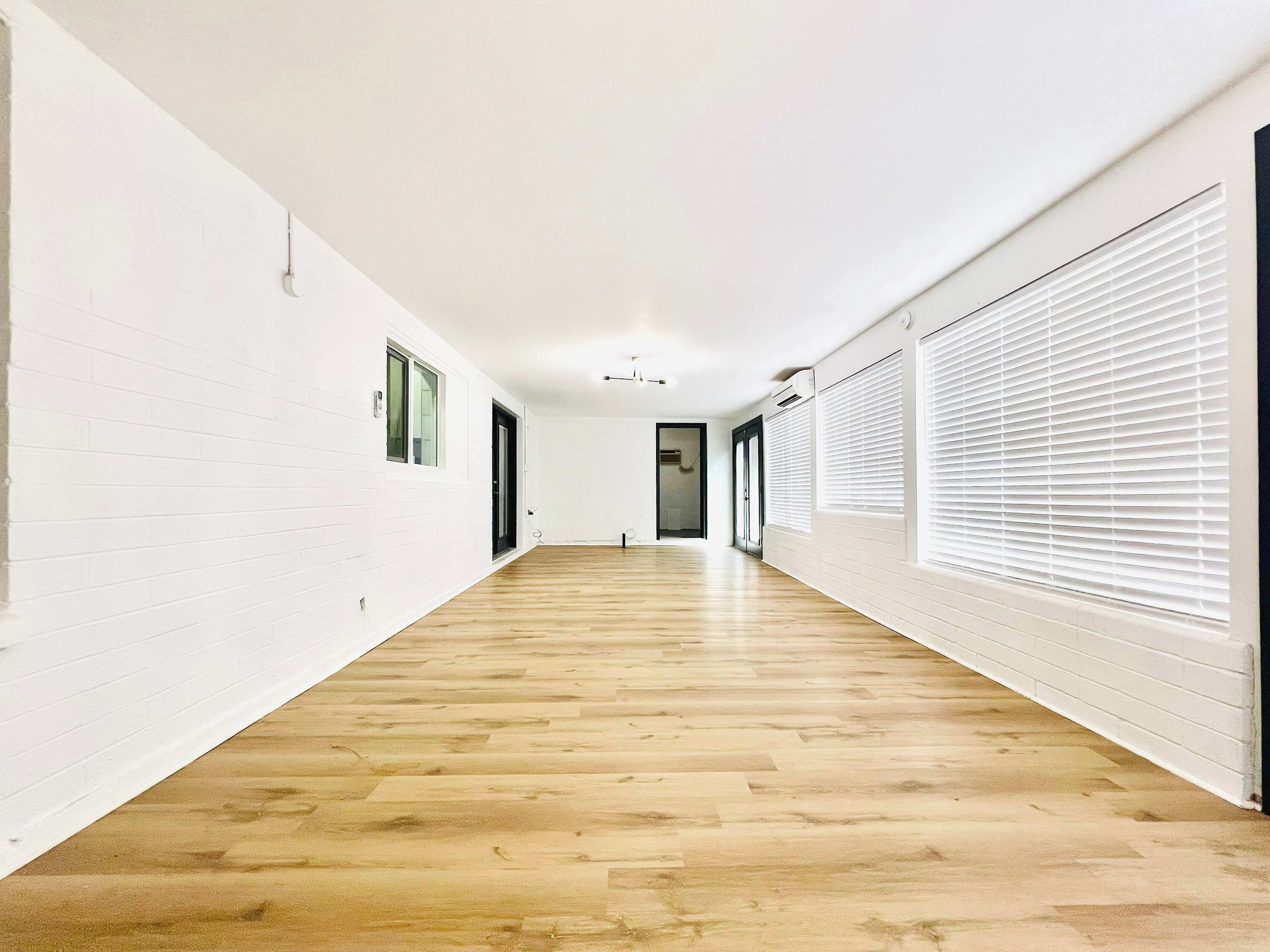 A long hallway with white brick walls, wooden flooring, and large windows with blinds.