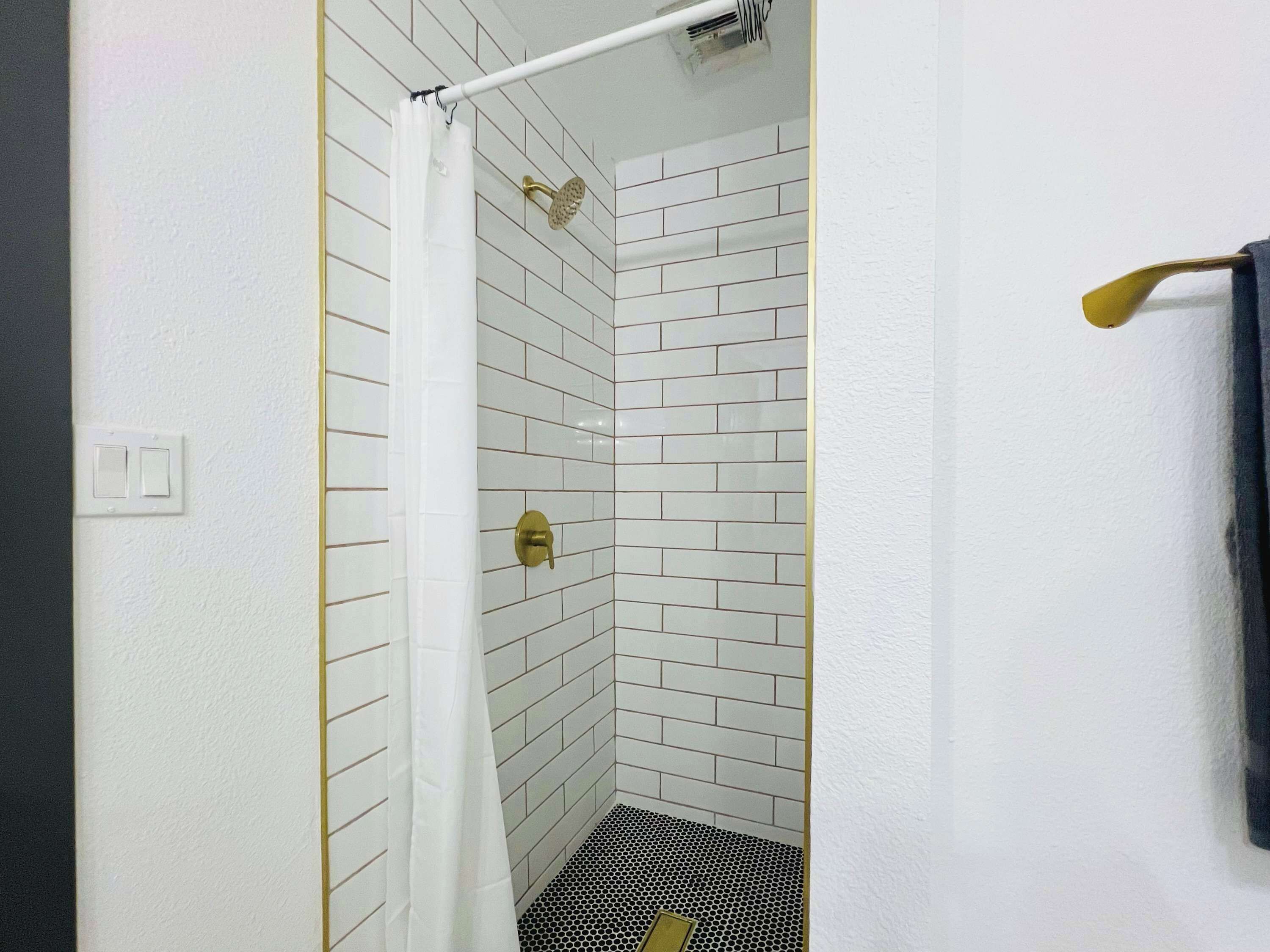 Une douche aux murs recouverts de carreaux blancs, avec une pomme de douche dorée et un rideau blanc.