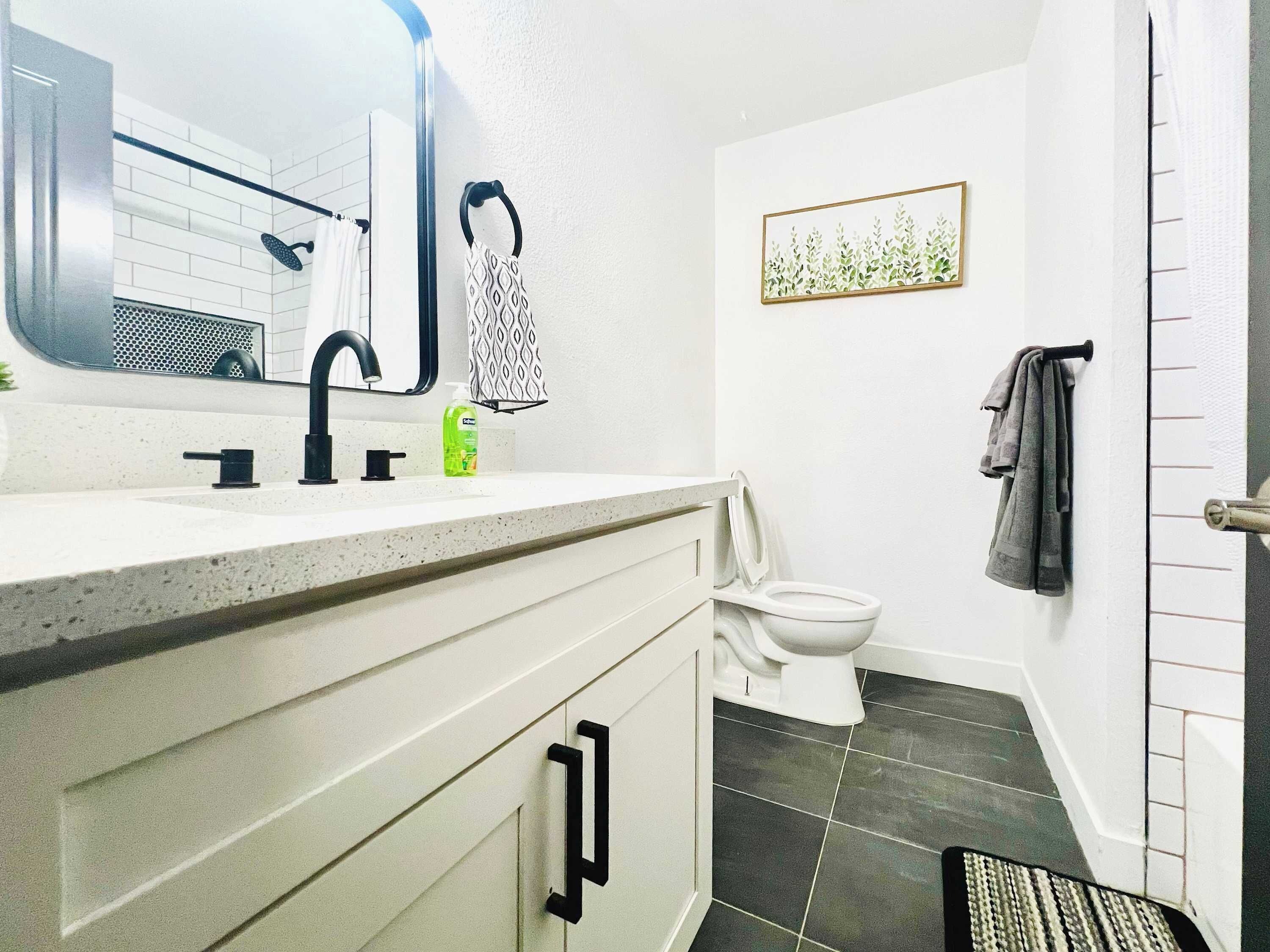 Een badkamer met een witte wastafel, een zwarte kraan en een toilet. Er is een handdoekhouder met een handdoek met patroon en een lijst met foto aan de muur.