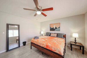 Un dormitorio con cama, ventilador de techo, mesita de noche con lámpara, un cuadro enmarcado en la pared y un espejo.