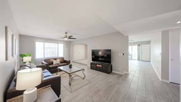 A spacious living room with a flat-screen TV, a ceiling fan, and a large window.